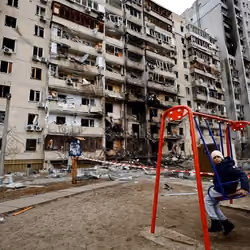 A child sits on a swing in front of a damaged residential building. 